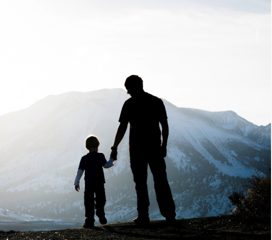 Silhouette of a father and child holding hands against a snow-covered mountain range at sunrise.