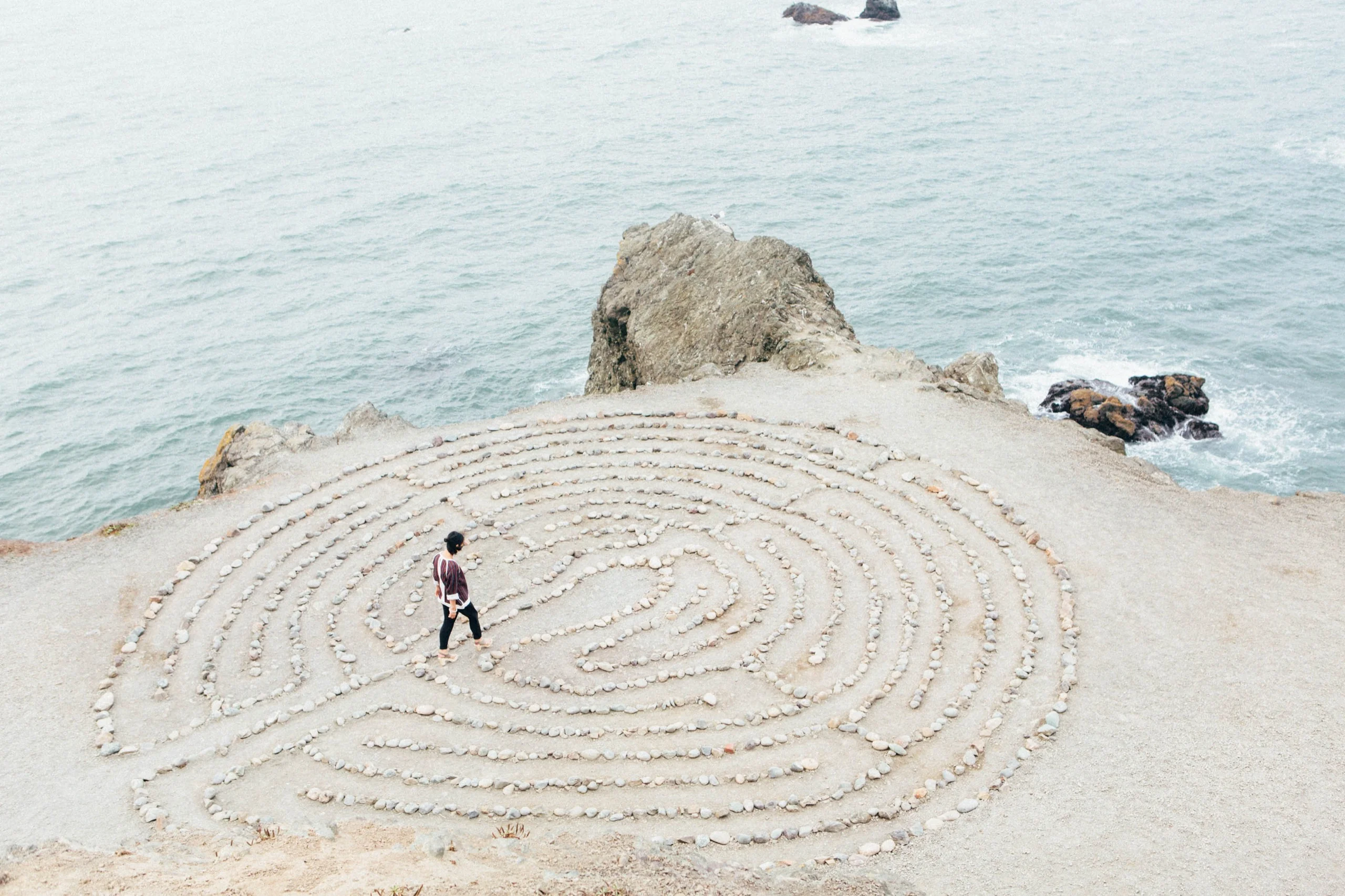 A person standing at the centre of a large stone labyrinth laid out on a headland above the sea